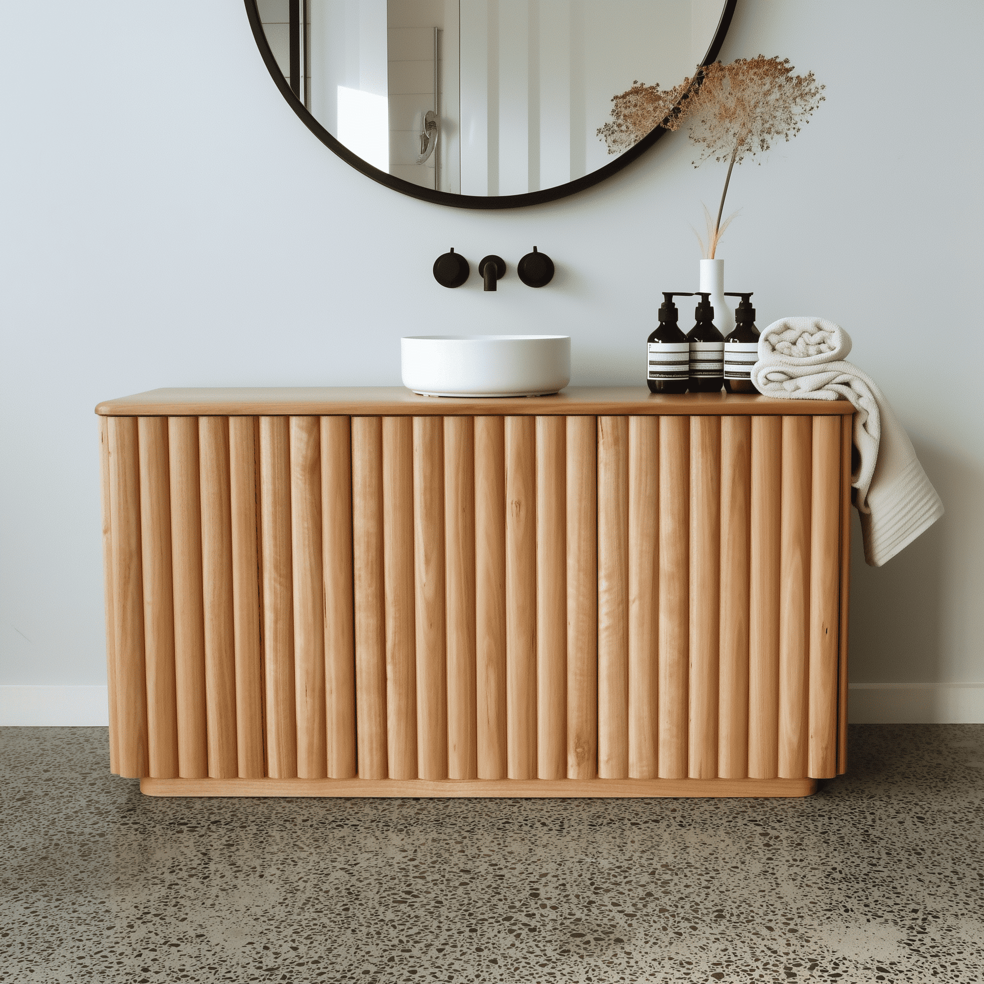 Oak bathroom vanity with round mirror, toiletries, and towel on a light gray wall background.