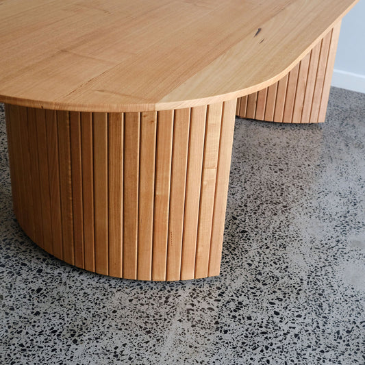 oak timber dining table with a ribbed cylindrical base on a speckled floor.