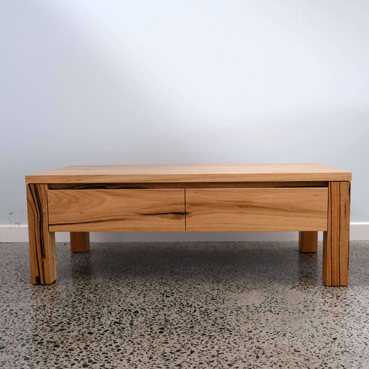 Wooden coffee table with a single drawer on a speckled floor against a white wall